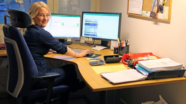 Blond kvinna med glasögon vid ett skrivbord med två dataskärmar.