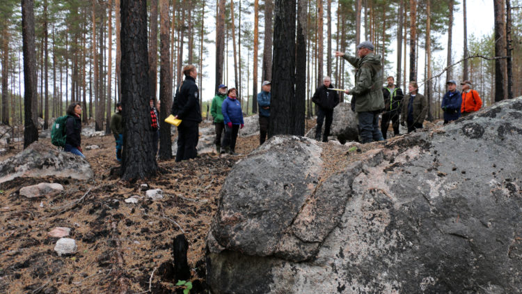 Ett tiotal människor står i skog som brunnit. I förunden en stor sten.