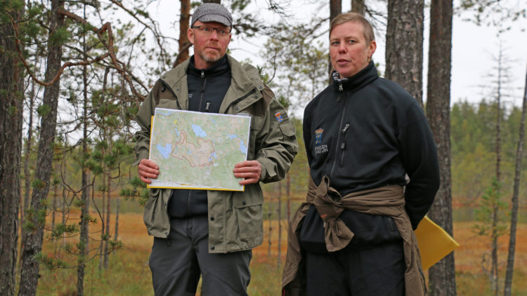 En man och en kvinna står framför tallar. Mannen visar upp en karta.