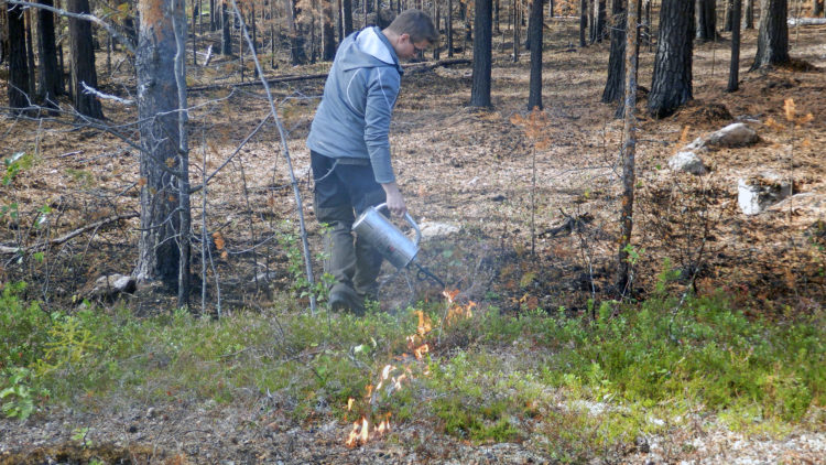 En man i grå jacka håller en silverfärgad tändkanna som droppar eld.