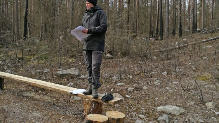 En kvinna står på en bänk i en skog. Hon har papper i handen.