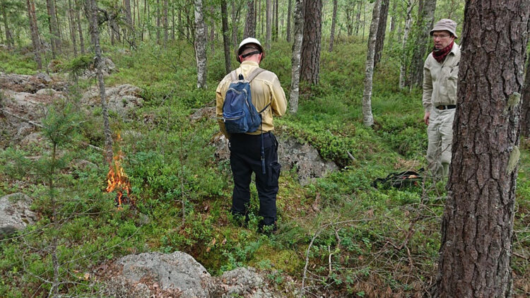 En man med svartabyxor, gul skjorta och blå ryggsäck står bredvid en liten eld.