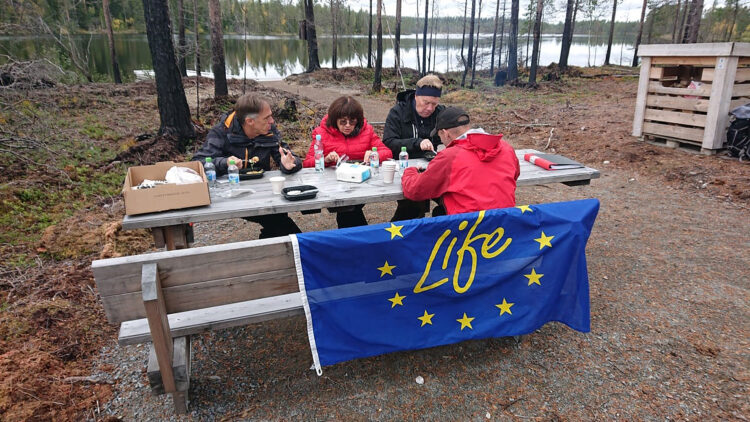 Fyra personer sitter på bänkar vid ett bord ute i naturen och äter.