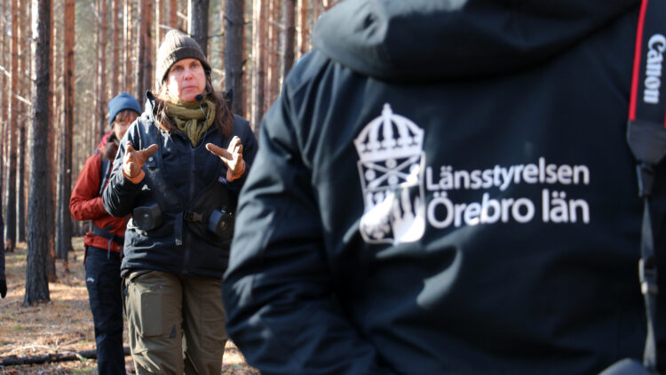 En kvinna i friluftskläder pratar. Närmast i bild en jacka med trycket Länsstyrelsen i Örebro län.