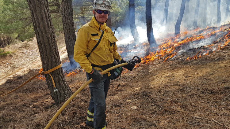 En man med gul skjorta och vit hjälp håller i en brandslang. han står framför mark som brinner med låga lågor.
