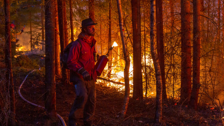 En man i röd skjorta och skägg går längs eldlinje som lyser upp honom i mörkret.